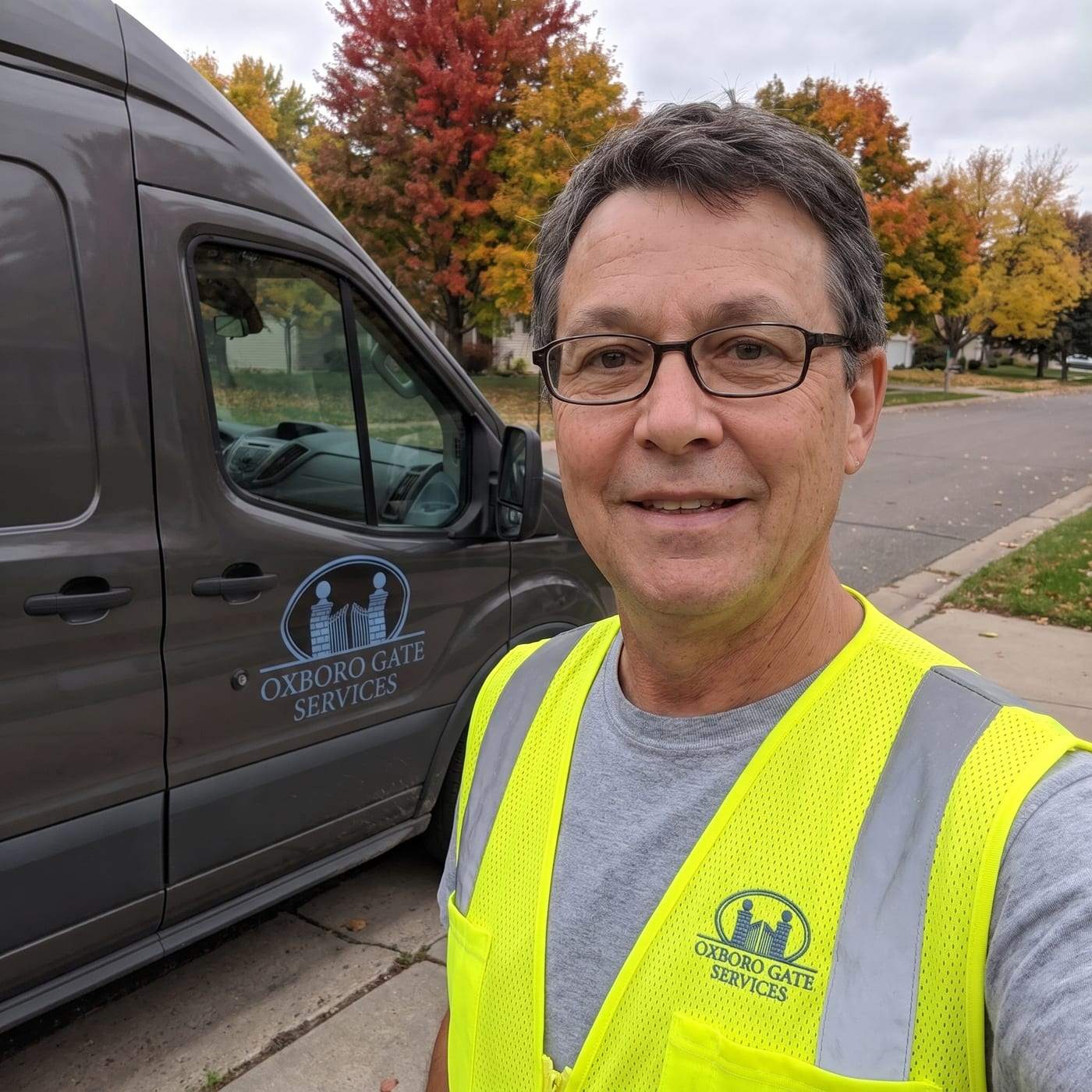 Gilbert Mckay in a hi-vis vest with the company logo, standing beside the charcoal work van at the curb