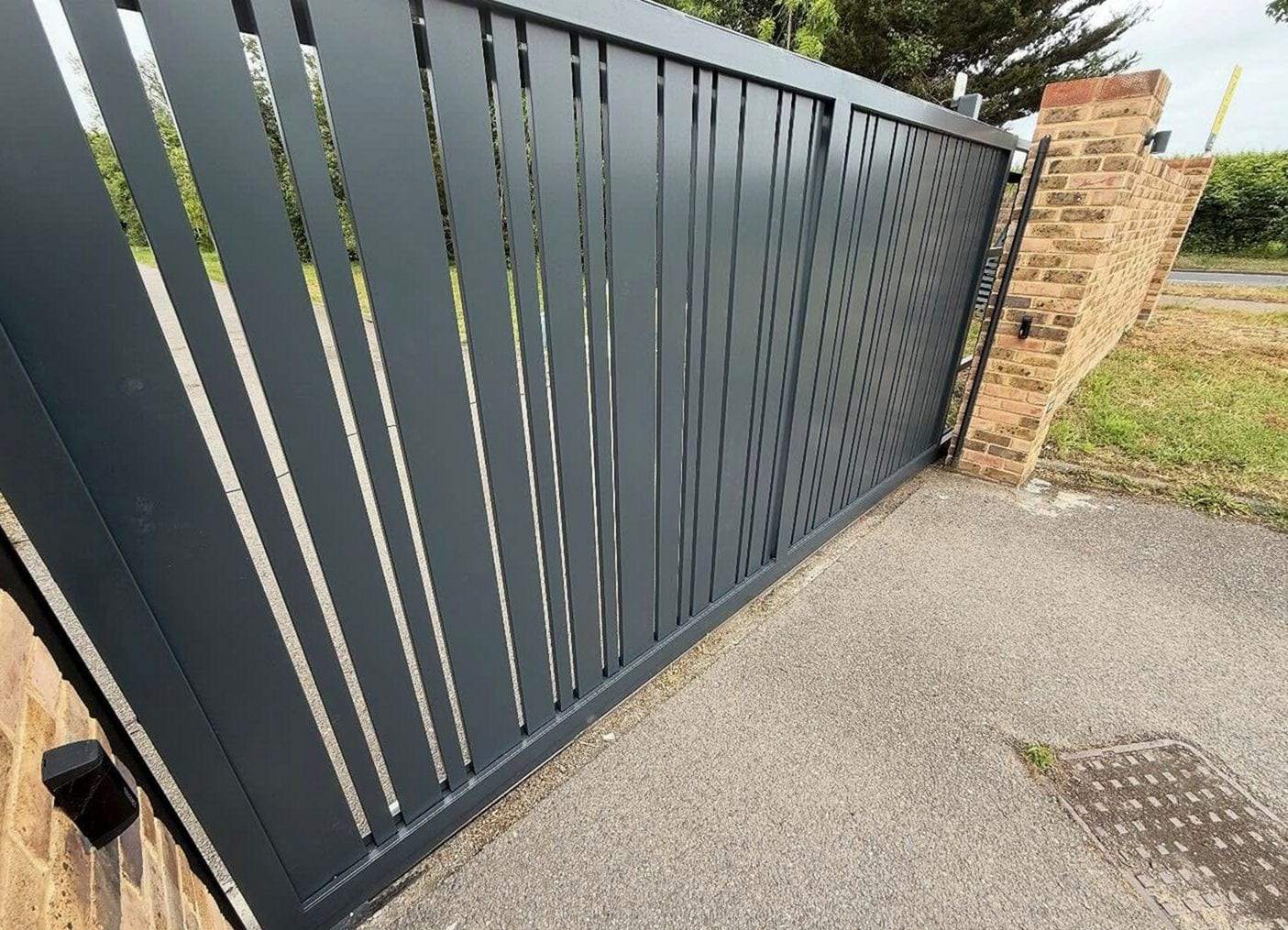 Charcoal slatted slide gate closed across a paved driveway beside a brick pier with a photo eye