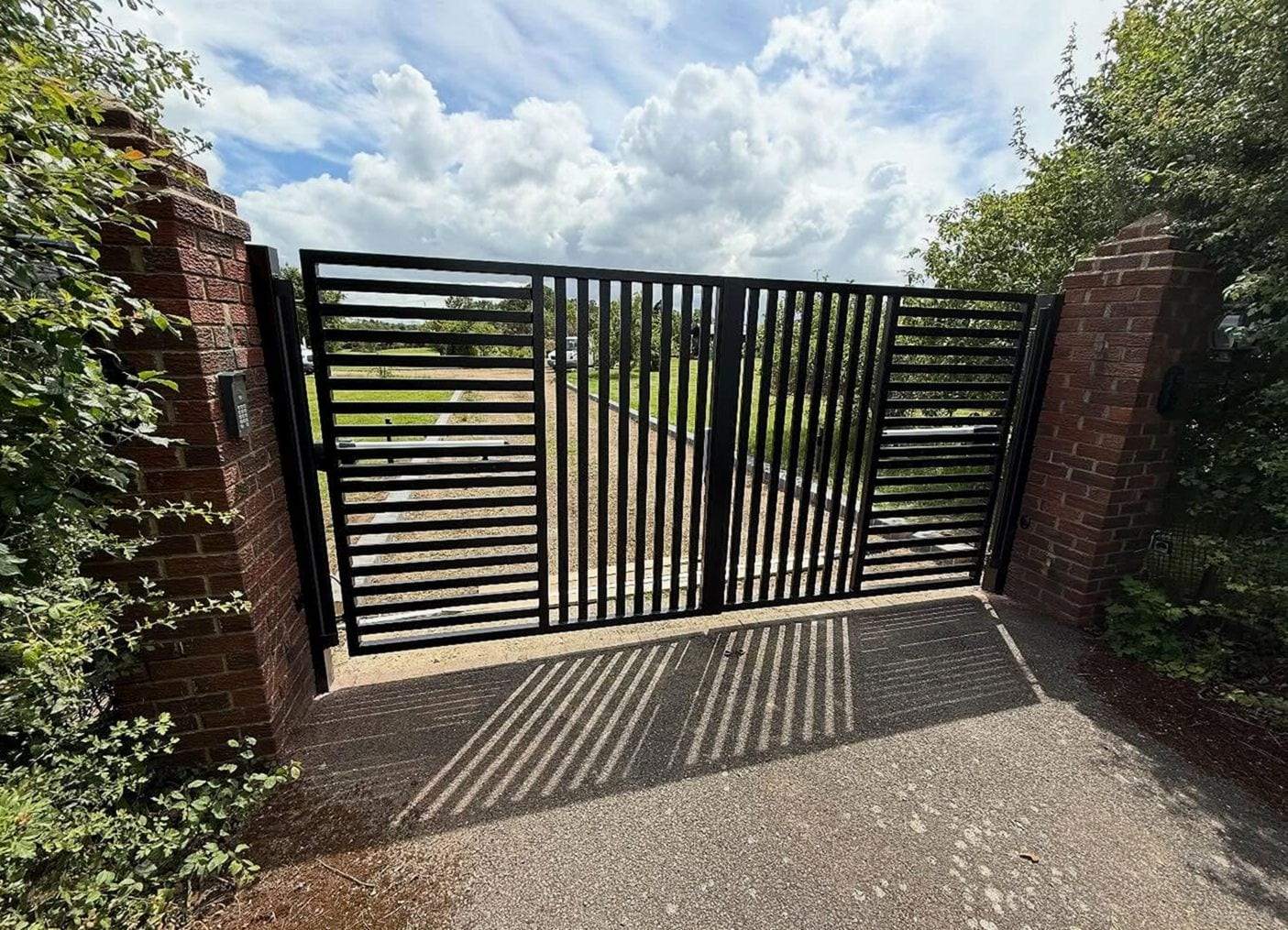 Black double swing driveway gate with mixed vertical and horizontal bars, hung between red-brick piers