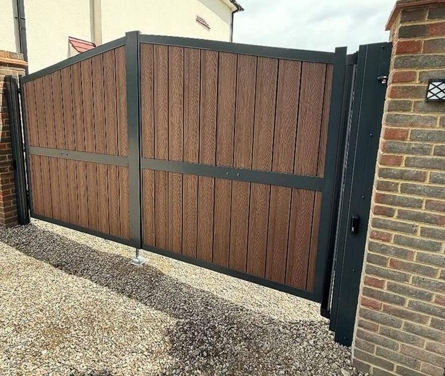 Wood-look composite double swing gate in a charcoal steel frame on a gravel drive beside a brick pillar