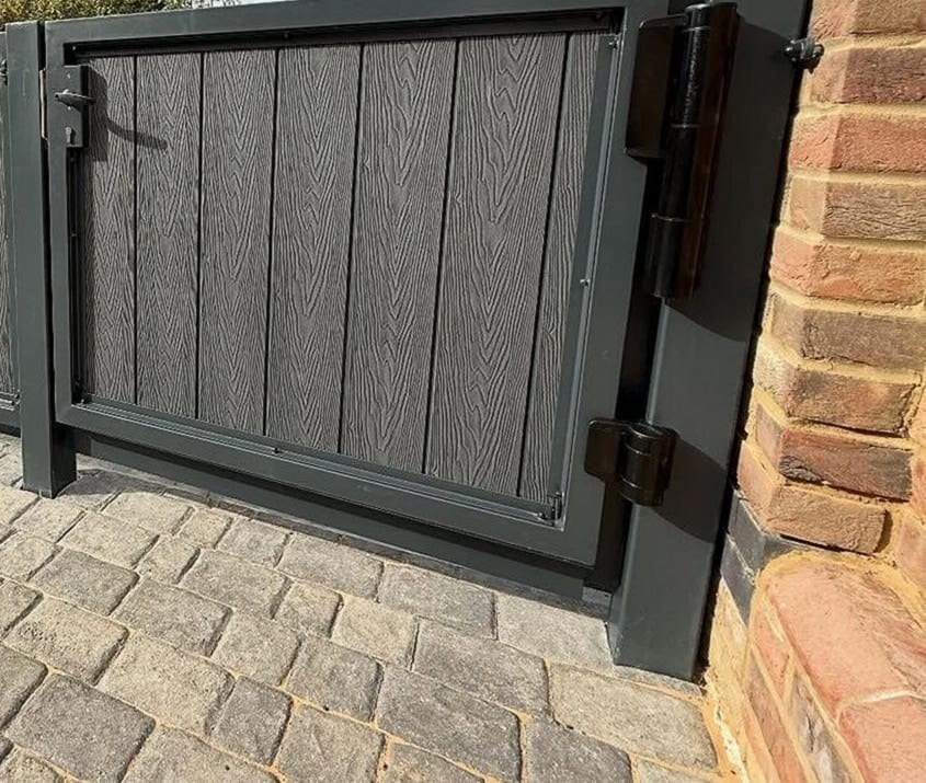 Gray wood-grain composite walk gate hung on heavy black hinges at a brick pier