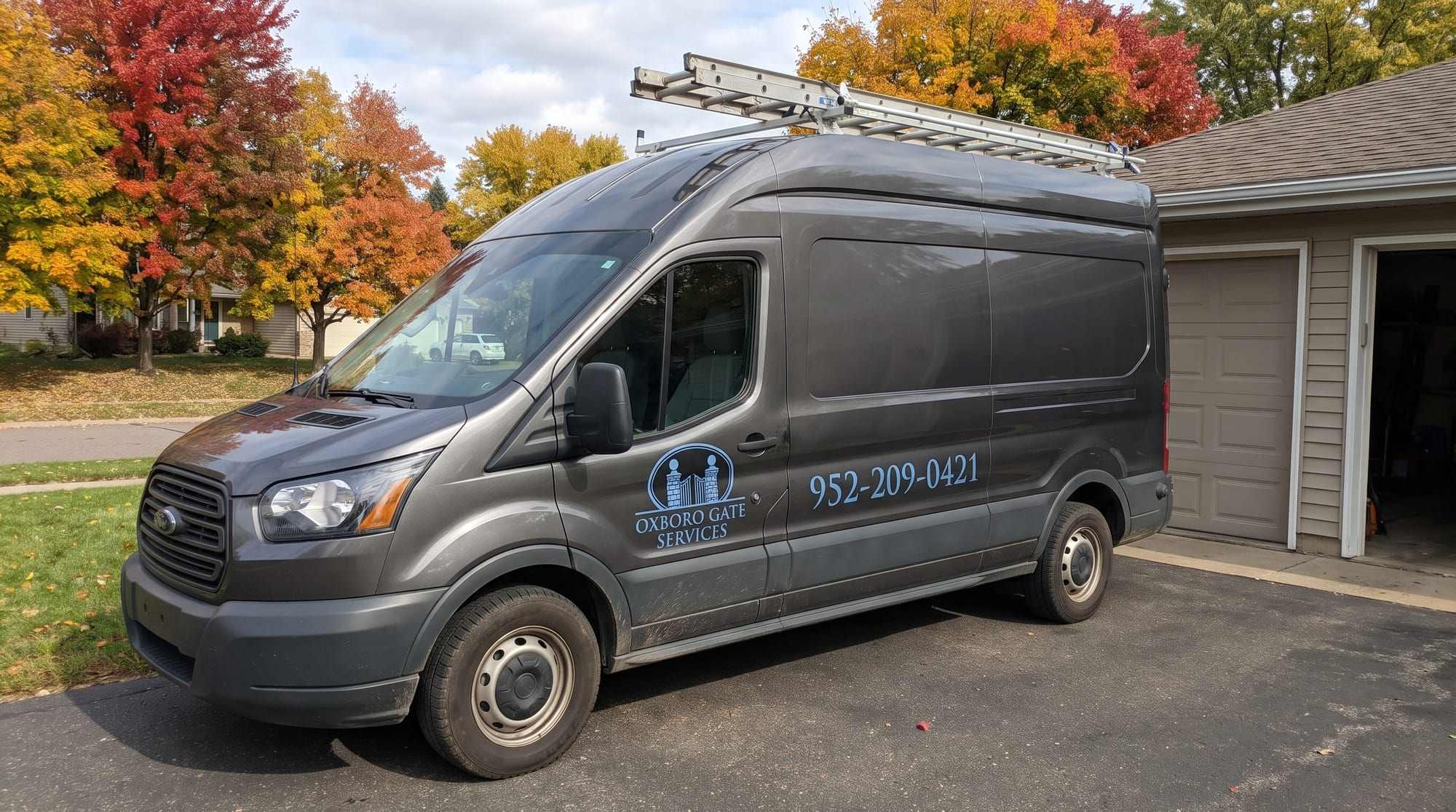Charcoal Ford Transit high-roof van with the Oxboro Gate Services logo on the front door and 952-209-0421 on the side, parked in a driveway under autumn maples